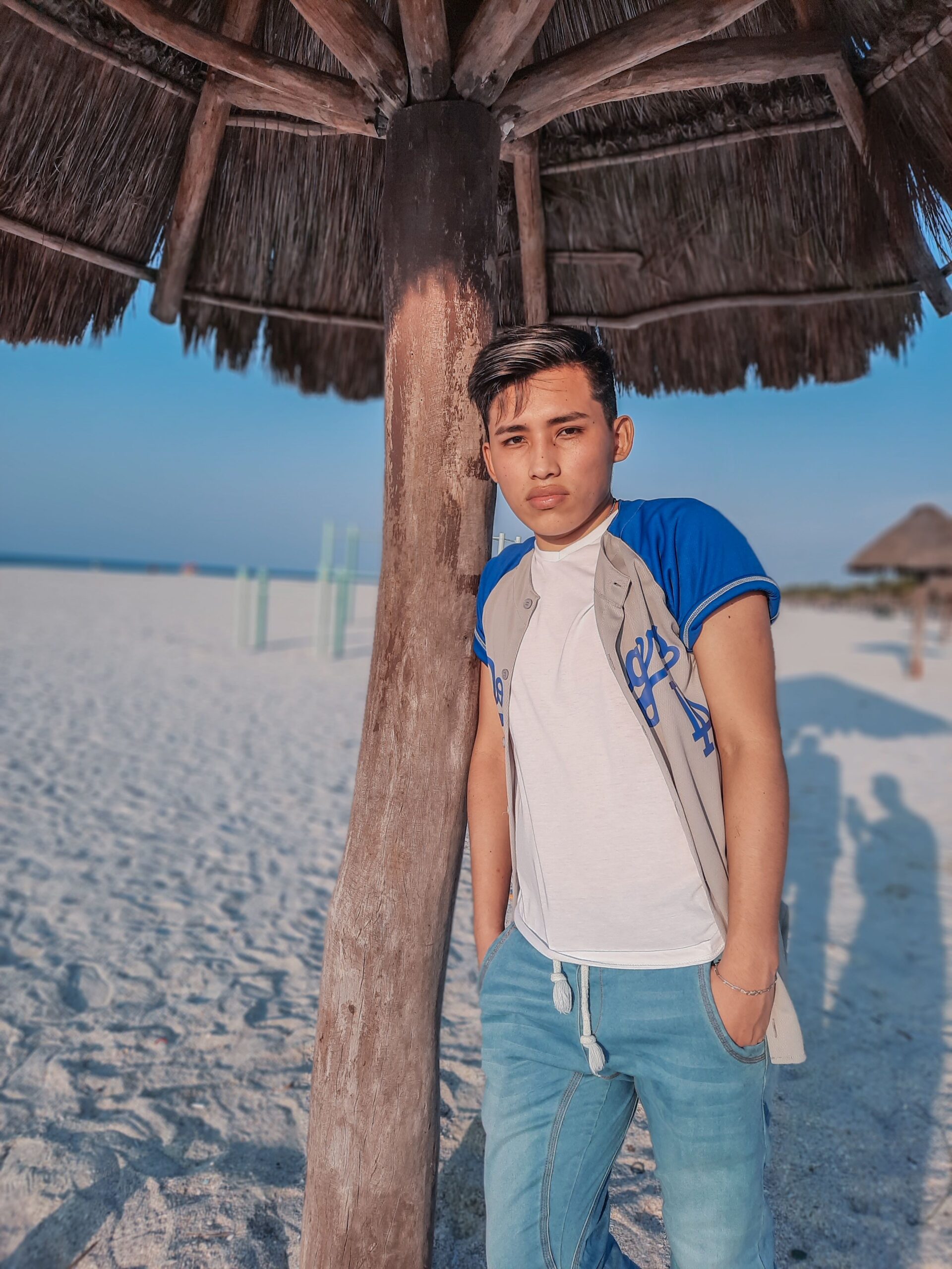 Young man in casual attire leaning on a beach umbrella under a clear sky.