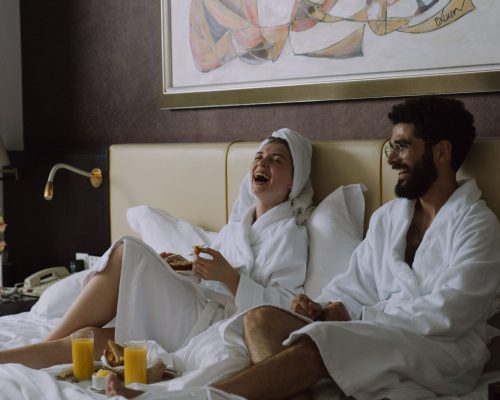 A joyful couple in bathrobes laughing while having breakfast in bed indoors.
